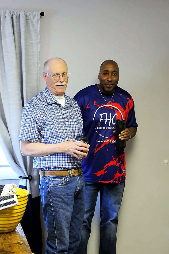 Two men stand side by side holding coffee cups, one wearing a plaid shirt and the other a blue Freedom House Church shirt that reads welcome home.