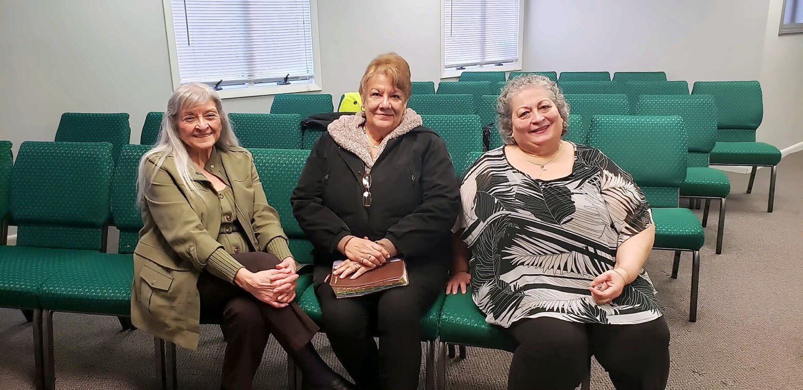 Three women sit together on green chairs in the Freedom House Church sanctuary, smiling at the camera, one holding a Bible.
