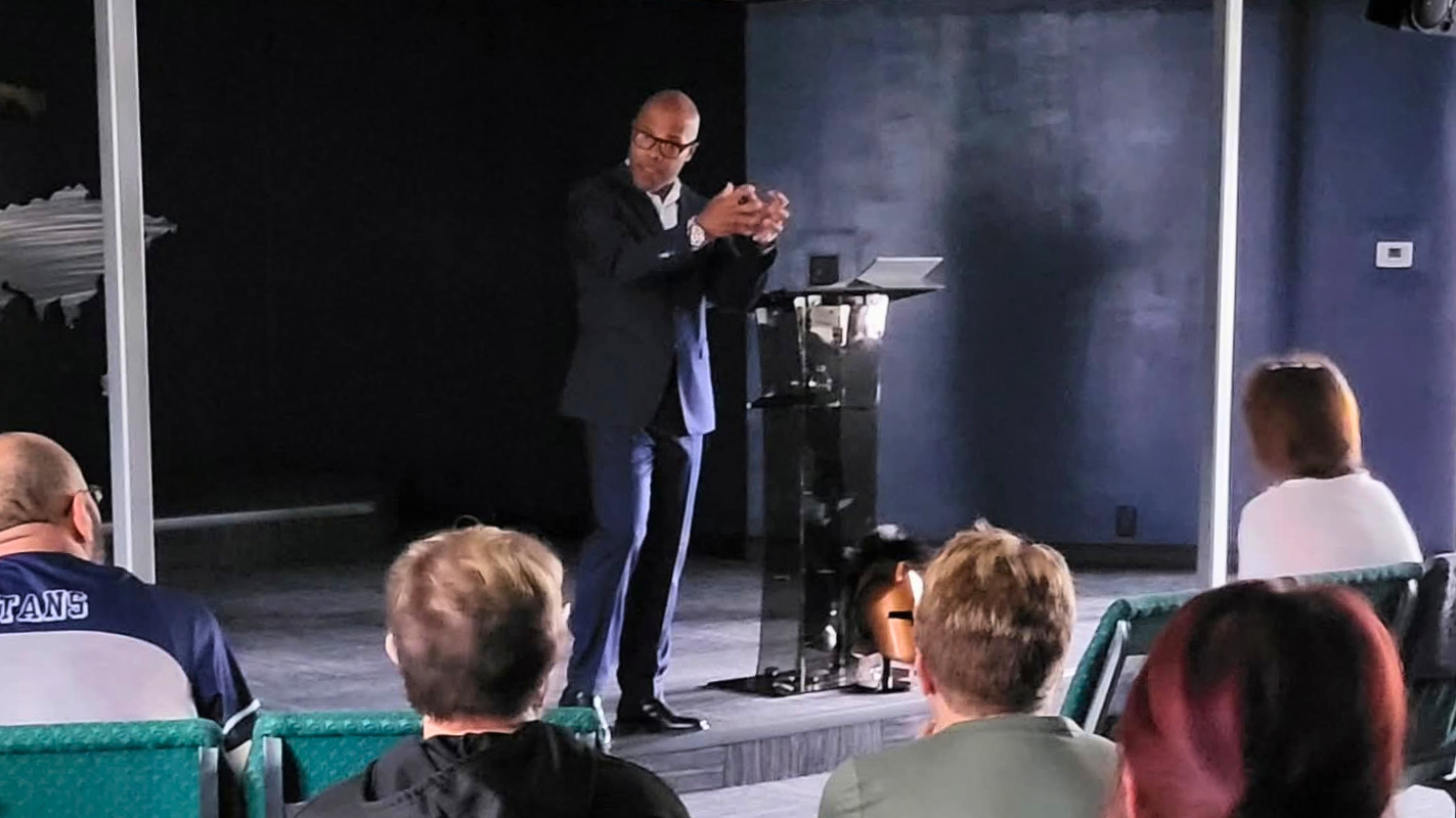 Pastor Jason, in a dark suit, speaks from the stage beside a clear podium while members of the congregation listen from their seats.